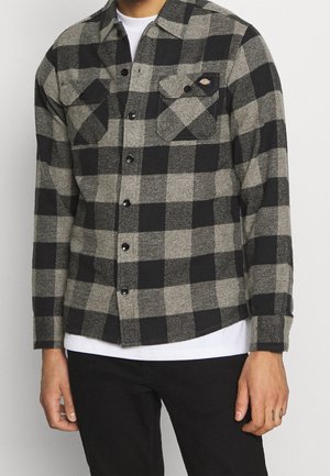 Mann trägt ein schwarz-grau kariertes Hemd mit Knöpfen über einem weißen T-Shirt und schwarzen Hosen, mit Taschen und einem kleinen Logo auf der Brust.