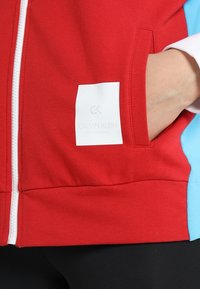 Giacca rossa con zip, dotata di una cerniera bianca, tasca laterale e un'etichetta bianca che mostra "CALVIN KLEIN PERFORMANCE". Accenti blu chiaro sulle maniche.
