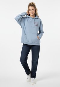 Felpa azzurra chiara con tasca frontale e ricamo a forma di cuore rosso, abbinata a jeans scuri e sneaker bianche. Tessuto morbido, vestibilità rilassata.