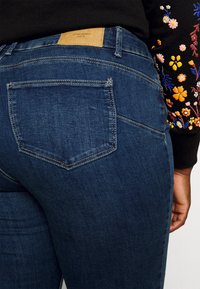 Dark blue denim jeans with a high waist, featuring a leather tag at the back and a back pocket with a curved seam detail.