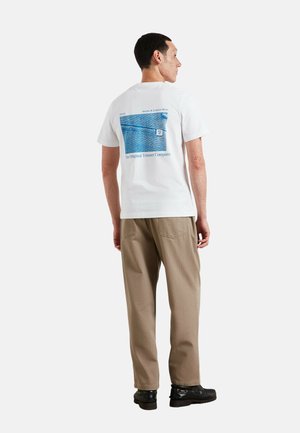 Mężczyzna ubrany w biały T-shirt z nadrukiem w stylu niebieskiego denimu oraz beżowe spodnie, stojący tyłem na jednolitym tle.