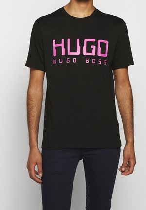 Man draagt een zwart T-shirt met korte mouwen met grote roze "HUGO" en kleinere "HUGO BOSS" tekst over de borst.