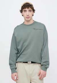 Sudadera verde de corte holgado, cuello redondo y puños acanalados. Tiene texto impreso en el lado izquierdo. Combinada con pantalones cortos beige.