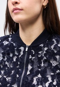 Κοντινό πλάνο γυναίκας που φορά χρυσούς κρίκους σκουλαρίκια και μπλε ναυτικό δαντελένιο σακάκι με ασημί φερμουάρ μπροστά και ριμπ ύφασμα στο γιακά.