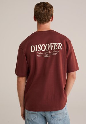 Bordeaux katoenen t-shirt met korte mouwen. Grote witte tekst op de rug leest "ONTDEK" en extra tekst in kleinere letters eronder.
