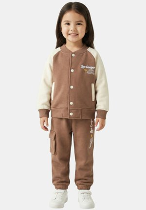 Jeune fille souriante, portant une veste boutonnée marron et crème assortie à un pantalon cargo marron et des baskets blanches.