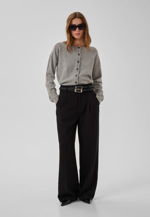 Mujer con pantalones negros de pierna ancha, cárdigan gris abotonado, cinturón negro, zapatos puntiagudos y grandes gafas de sol negras, de pie con las manos en los bolsillos.