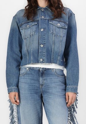 Femme portant une veste en denim bleue courte boutonnée et un jean bleu taille haute avec des détails à franges sur les côtés.