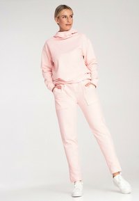 Jasnoróżowa bluza z kapturem i długimi rękawami, zestawiona z dopasowanymi spodniami z przodu z kieszeniami, noszona z białymi sneakersami.