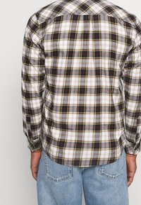 Camicia flanella a scacchi in nero, bianco, rosso e grigio. Maniche lunghe con polsini abbottonati. Orlo arrotondato e dettaglio della spalla sagomato. Indossata con jeans blu.
