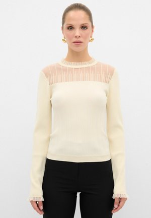 Maglione a coste crema con maniche lunghe e pannello trasparente in rete nella parte superiore, arricciato sui polsini, abbinato a pantaloni neri aderenti.