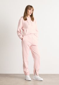 Ροζ φούτερ με κορδόνι και μπροστινή τσέπη, σε συνδυασμό με αντίστοιχες φόρμες που διαθέτουν ριμπ μανσέτες και φερμουάρ στους αστραγάλους. Λευκά αθλητικά παπούτσια.