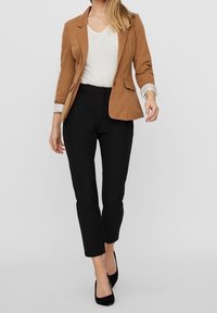 Hnědý blazer přes bílý top, v kombinaci s černými kalhotami. Blazer má dvě kapsy, slim fit a ohrnuté rukávy. Obuví jsou černé lodičky.