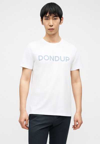 Νεαρός άνδρας που φοράει λευκό κοντομάνικο μπλουζάκι με μπλε κείμενο "DONDUP" και σκούρο παντελόνι, στέκεται μπροστά σε έναν απλό φωτεινό φόντο.
