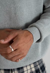 Ruke osobe koja nosi dva srebrna prstena, svijetlosivu duksericu i hlače s uzorkom crno-bijelog kariranog dizajna, s prstima nježno spojeno.