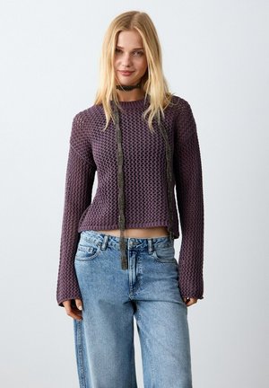 Donna che indossa un maglione viola a maglia con maniche lunghe, jeans blu a vita alta e una collana nera con catena su sfondo semplice.