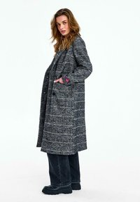 Žena s dlhými vlnitými vlasmi v sivej kárované dlhej vrstve, čiernych nohaviciach s širokými nohavicami a čiernych robustných topánkach, stojaca bokom s jednou rukou v vrecku.