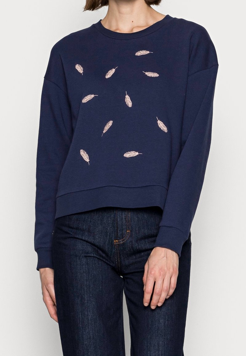Femme portant un sweat bleu marine avec des motifs de plumes rose clair dispersés sur le devant, associé à un jean bleu foncé.