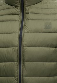 Veste matelassée vert olive avec fermeture éclair noire et petite étiquette carrée portant l'inscription « BOSS » sur la poitrine.