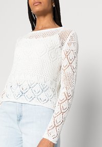 Pull blanc en tricot avec un design texturé ajouré, manches longues et ourlet court, associé à un jean bleu clair.
