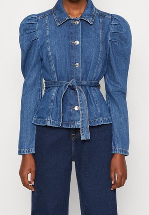 Denimjakke med puffede skuldre, sølvknapper foran, bundet bælte i taljen, båret med mørkeblå denimjeans på en person.