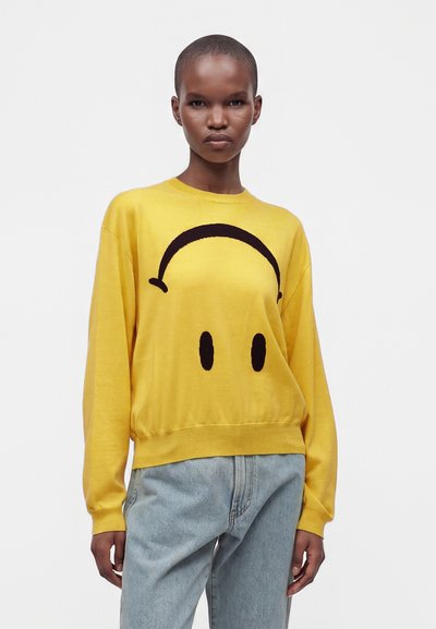 Personne portant un pull jaune avec un grand motif smiley noir et un jean bleu clair sur un fond blanc uni.