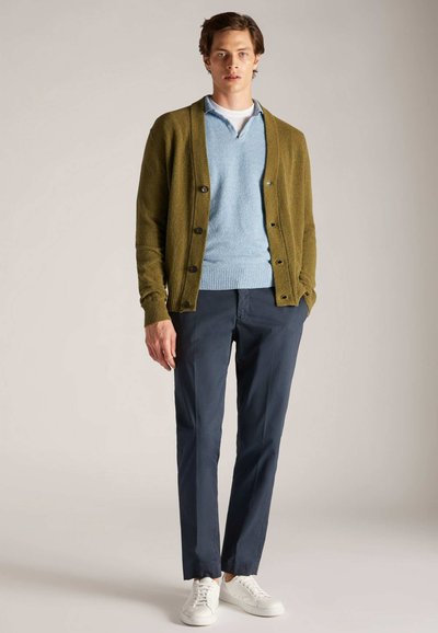 Un joven se presenta con confianza, vistiendo un cárdigan verde sobre un suéter azul claro y pantalones azul marino, completando el atuendo con zapatillas blancas.