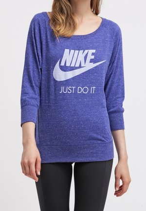 Femme portant un sweat-shirt Nike bleu à manches longues avec le slogan « Just Do It » imprimé sur le devant, associé à un legging noir.