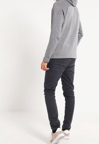 Grå hoodie med texturerad tyg, uppsatt mot svarta slim-fit byxor. Ljusfärgade sneakers fulländar outfiten. Bakifrån, enfärgad bakgrund.
