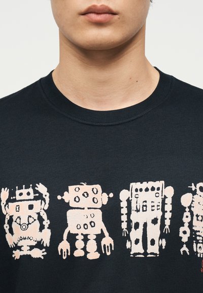 Κοντινό πλάνο ενός ατόμου που φοράει μαύρο T-shirt με τέσσερις στυλιζαρισμένες, αφηρημένες εικονογραφήσεις ρομπότ σε ανοιχτό ροζ χρώμα στο ύψος του στήθους.