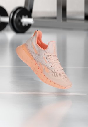 Αθλητικό παπούτσι σε ανοιχτό ροζ με κοραλί λεπτομέρειες, να αιωρείται πάνω από μια αντανακλαστική γυμναστηριακή επιφάνεια, με θολά όργανα άρσης βαρών στο φόντο.
