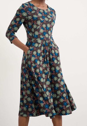 Vrouw draagt een midijurk met driekwart mouwen, voorzien van groene en blauwe bloemenpatronen, met handen in de zijzakken.