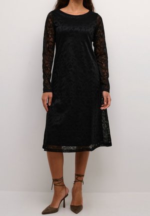 Kobieta ubrana w czarną koronkową sukienkę do kolan z długimi rękawami i okrągłym dekoltem, w połączeniu z brązowymi, spiczastymi szpilkami z paskami wokół kostek.