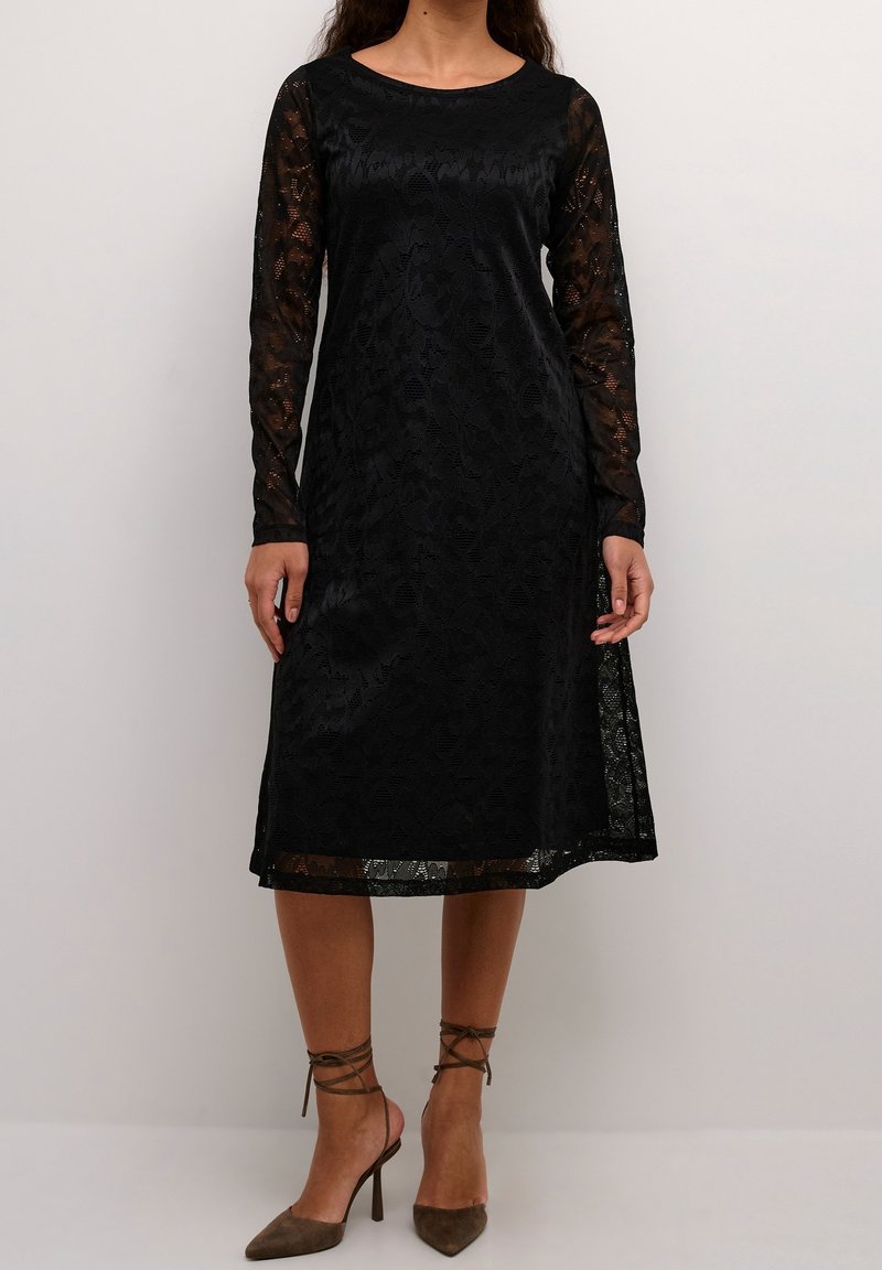 Femme portant une robe en dentelle noire mi-longue avec manches longues et encolure ronde, associée à des escarpins marron à bout pointu avec brides aux chevilles.
