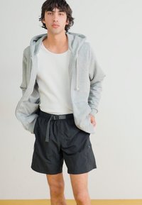 Pelēka kapuce ar rāvējslēdzēju, balta ribota topa krekliņa un melni šorti ar kabatām. Ikdienas, vieglas auduma. Modelis stāv pret vienkāršu fona.