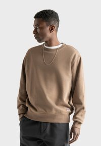 Bruine katoenen sweatshirt met geribbelde kraag en manchetten, voorzien van een klein label aan de onderzoom, gedragen over een wit shirt en gecombineerd met donkere broek.
