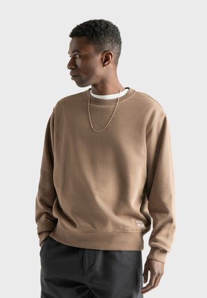Bruine katoenen sweatshirt met geribbelde kraag en manchetten, voorzien van een klein label aan de onderzoom, gedragen over een wit shirt en gecombineerd met donkere broek.