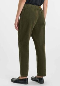 Persoon in olijfgroene ribfluweel broek met elastische tailleband en zwarte instappers, staande tegen een effen lichte achtergrond.