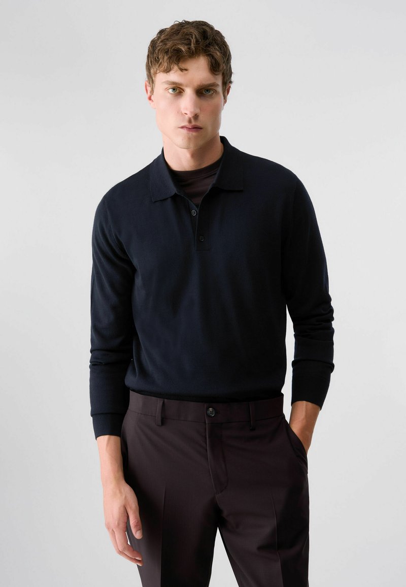 Jeune homme aux cheveux ondulés portant un polo noir à manches longues et un pantalon foncé, debout avec une main dans la poche devant un fond uni.