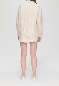 Camisa de manga larga de color beige claro con finas rayas verticales, combinada con pantalones cortos a juego, llevada con zapatos blancos con detalles metálicos.