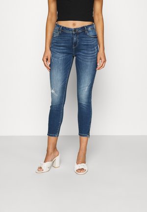 Kvinde iført mid-rise, let slidte cropped blå jeans, sort cropped top og hvide hælede sandaler med flettede remme, stående på gråt gulv.