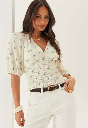 Vrouw met lang donker haar die een crèmekleurige gesmockte blouse met blauwe bloemenprint draagt, witte broek, donkere riem, gouden ketting en sieraden.