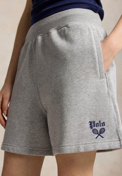 Gros plan d'une personne portant un short de survêtement gris avec une ceinture élastique et un logo "Polo" brodé avec deux raquettes de tennis.