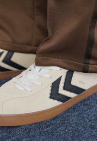 Beżowe zamszowe sneakersy z czarnymi zygzakowymi detalami, białymi sznurowadłami i brązową gumową podeszwą; połączone z brązowymi spodniami sportowymi. Niebieskie tło wykładziny.