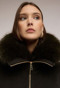 Mörk ullrock med en stor, plysj grön pälskrage, guldblixtlås och diskreta dragkedjedetaljer. Tyget verkar slätt med en strukturerad design.