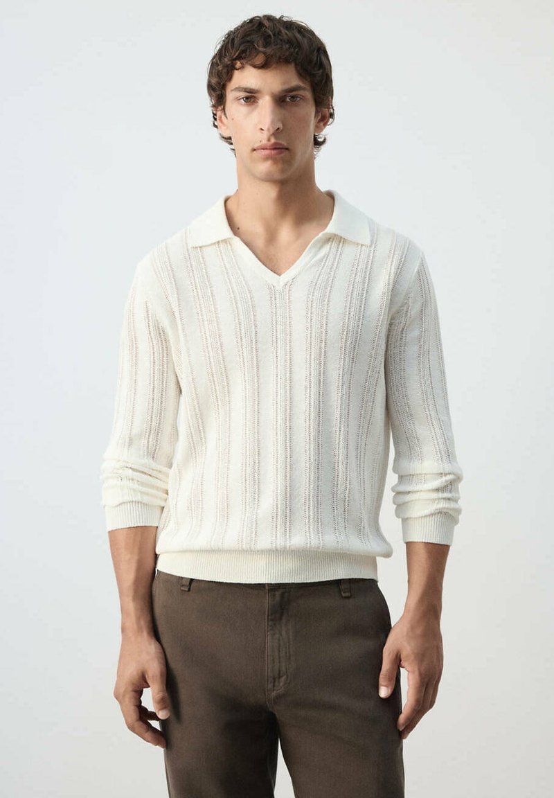 Jeune homme aux cheveux bouclés portant un pull blanc texturé à manches longues et un pantalon marron, debout devant un fond clair et uni.