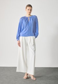 Lys blå, stripete bluse med nøkkelhull-hals og rynkede elastiske mansjetter, parret med hvite brede bukser og lyse sandaler.