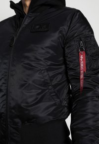 Giacca bomber nera in nylon lucido, con zip, polsini a costine, tasche laterali, logo a patch e un'etichetta rossa "RIMUOVI PRIMA DEL VOLO".