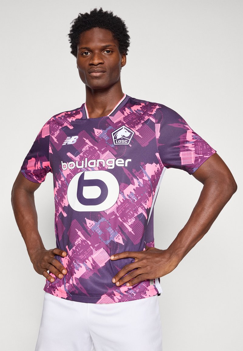 Maglietta da calcio viola a maniche corte con motivi geometrici rosa, dotata di colletto rotondo, dettagli bianchi e un grande logo bianco al centro.