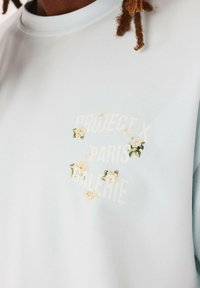 Sudadera de color azul claro con un diseño floral que presenta flores blancas y el texto "PROJECT X PARIS GALERIE" en una fuente sutil. Tela de algodón suave.